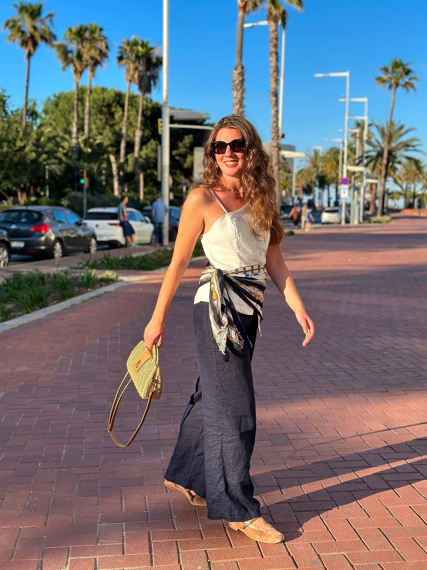 Miruna walking along a palm-lined promenade in Barcelona in linen trousers, a silk scarf tied at the waist.