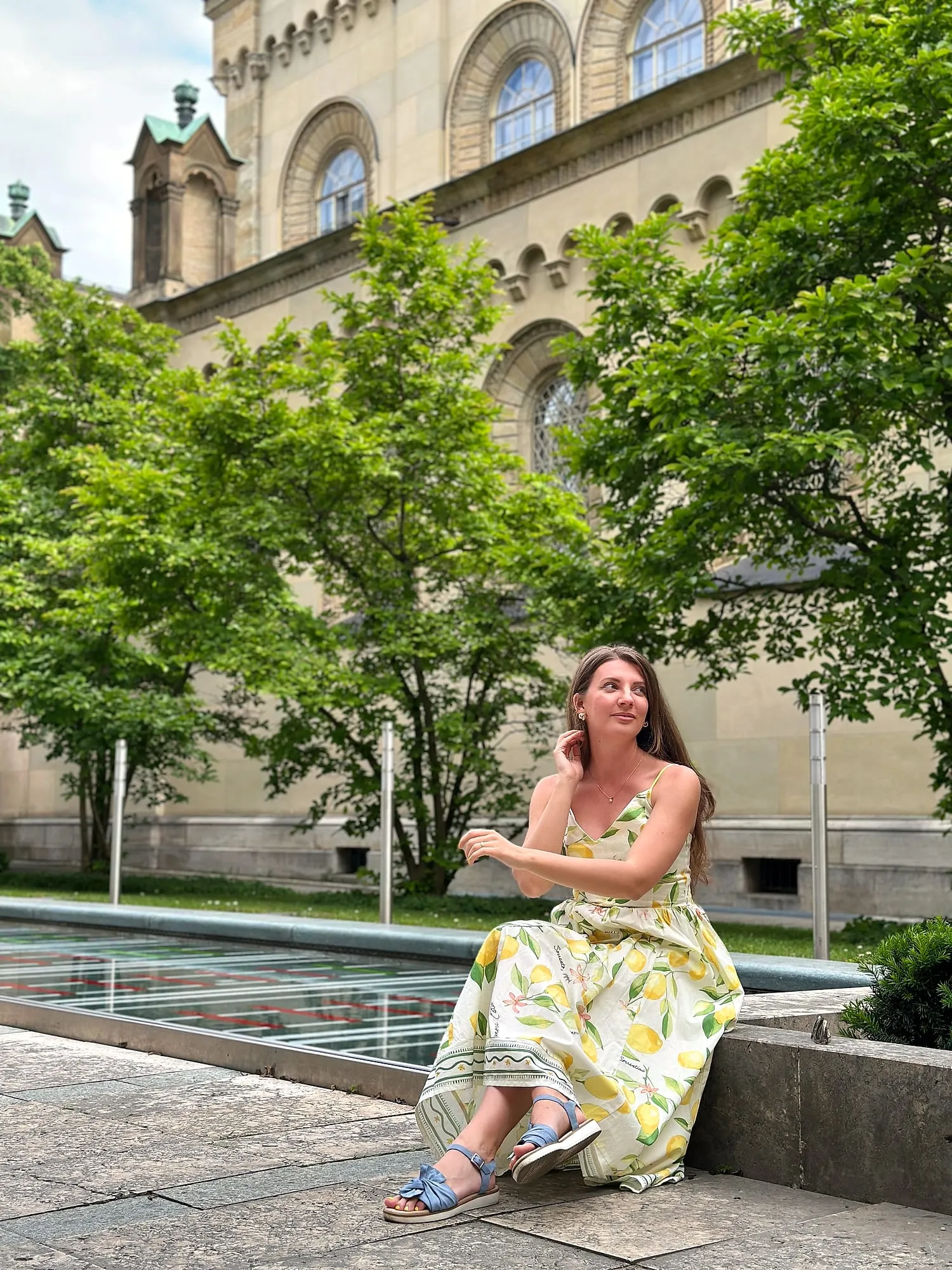 Miruna in a lemon-print summer dress, sitting at the edge of a fountain in the Munich Residenz garden.