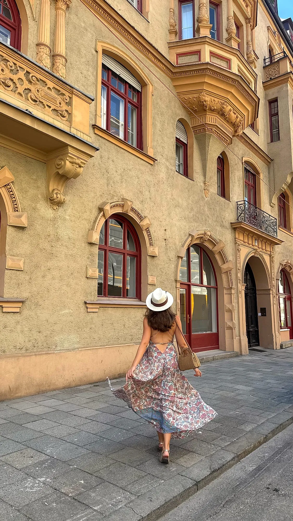 Miruna in a summer dress and straw hat walking past an ornate old-town facade in Munich.