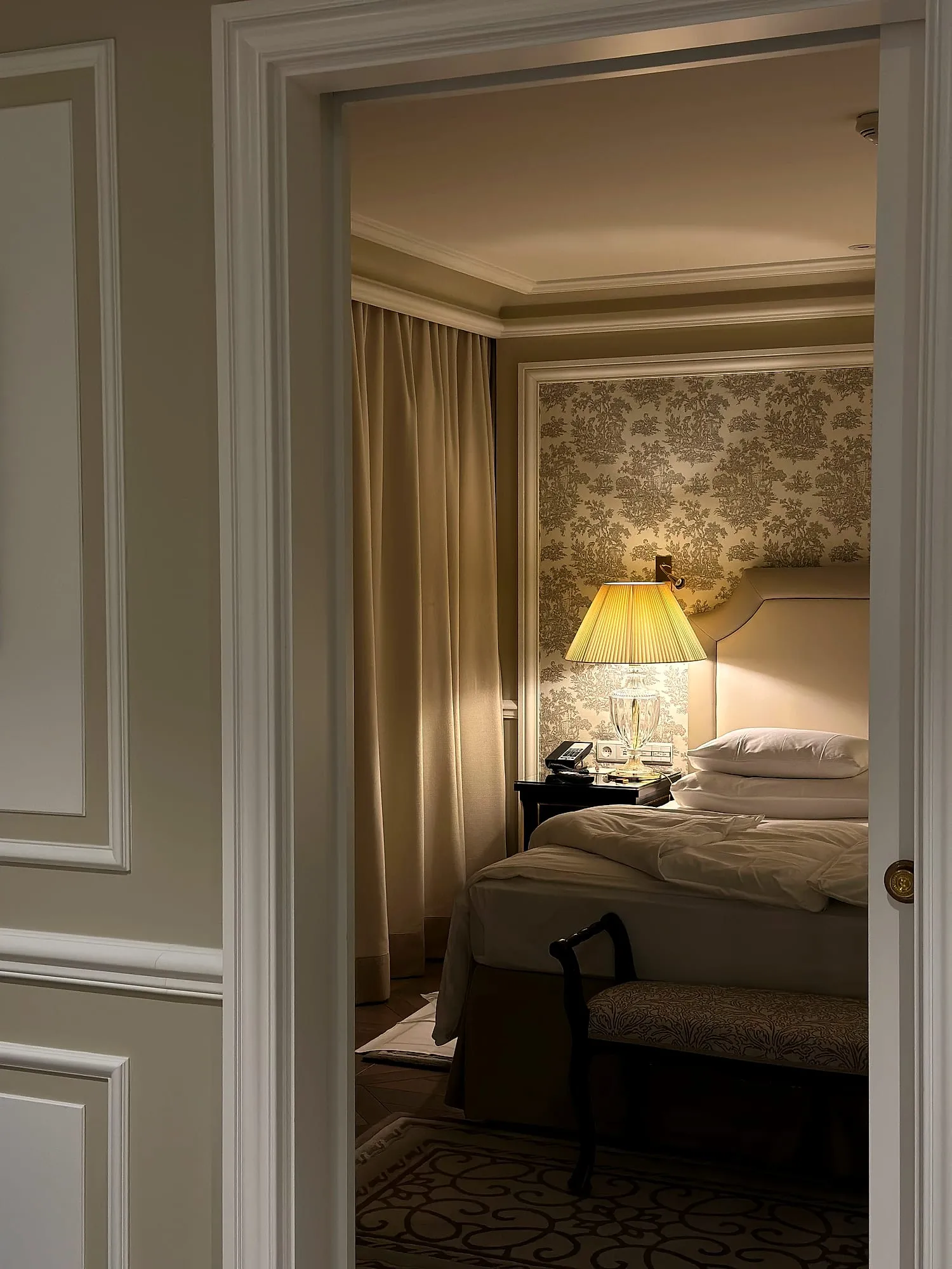 View into a suite at Hotel Sacher Salzburg, a bedside lamp glowing against toile wallpaper.