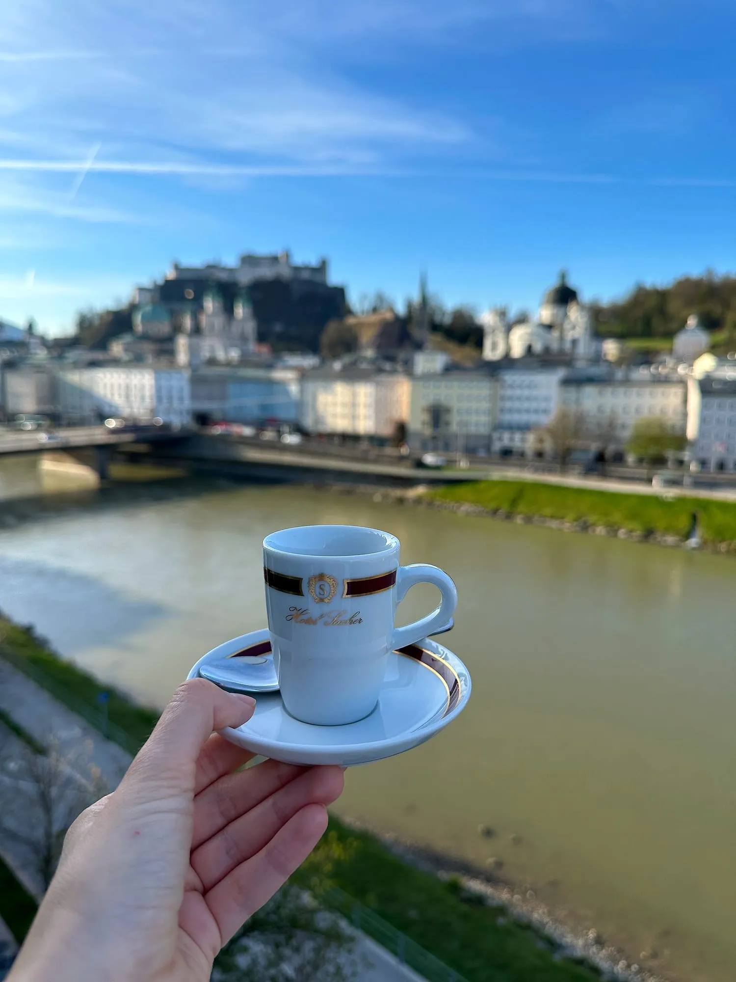 A Hotel Sacher espresso cup held up above the Salzach, the old town and the fortress of Salzburg behind.