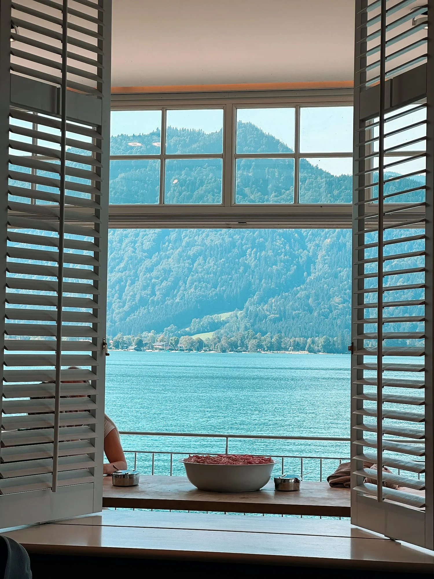 View through open white shutters across the Tegernsee to wooded mountains.