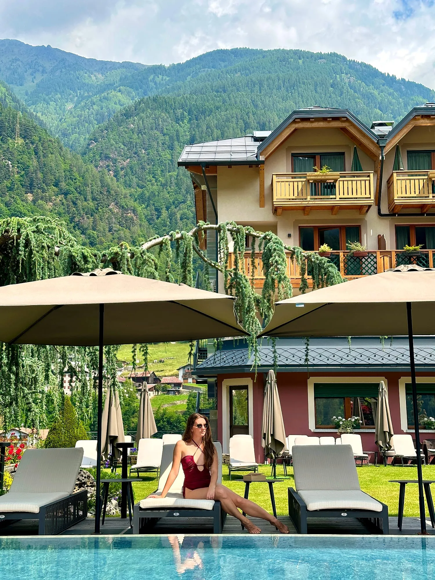 Miruna on a lounger at the pool of Tevini Charming Hotel, the house and the green mountains of Val di Sole behind.