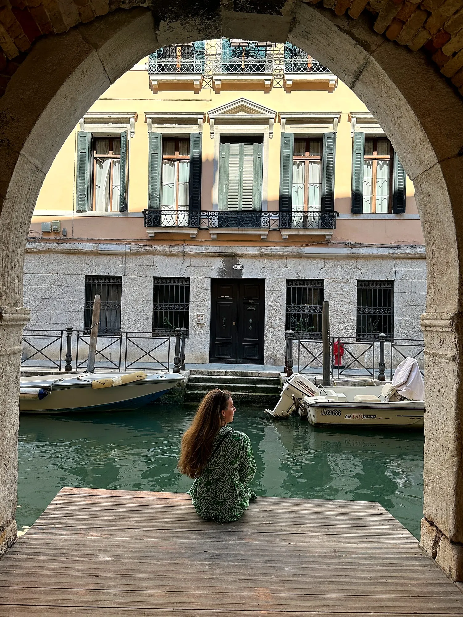 Miruna sitting on a wooden jetty beneath a stone archway, looking out over a quiet canal in Venice.